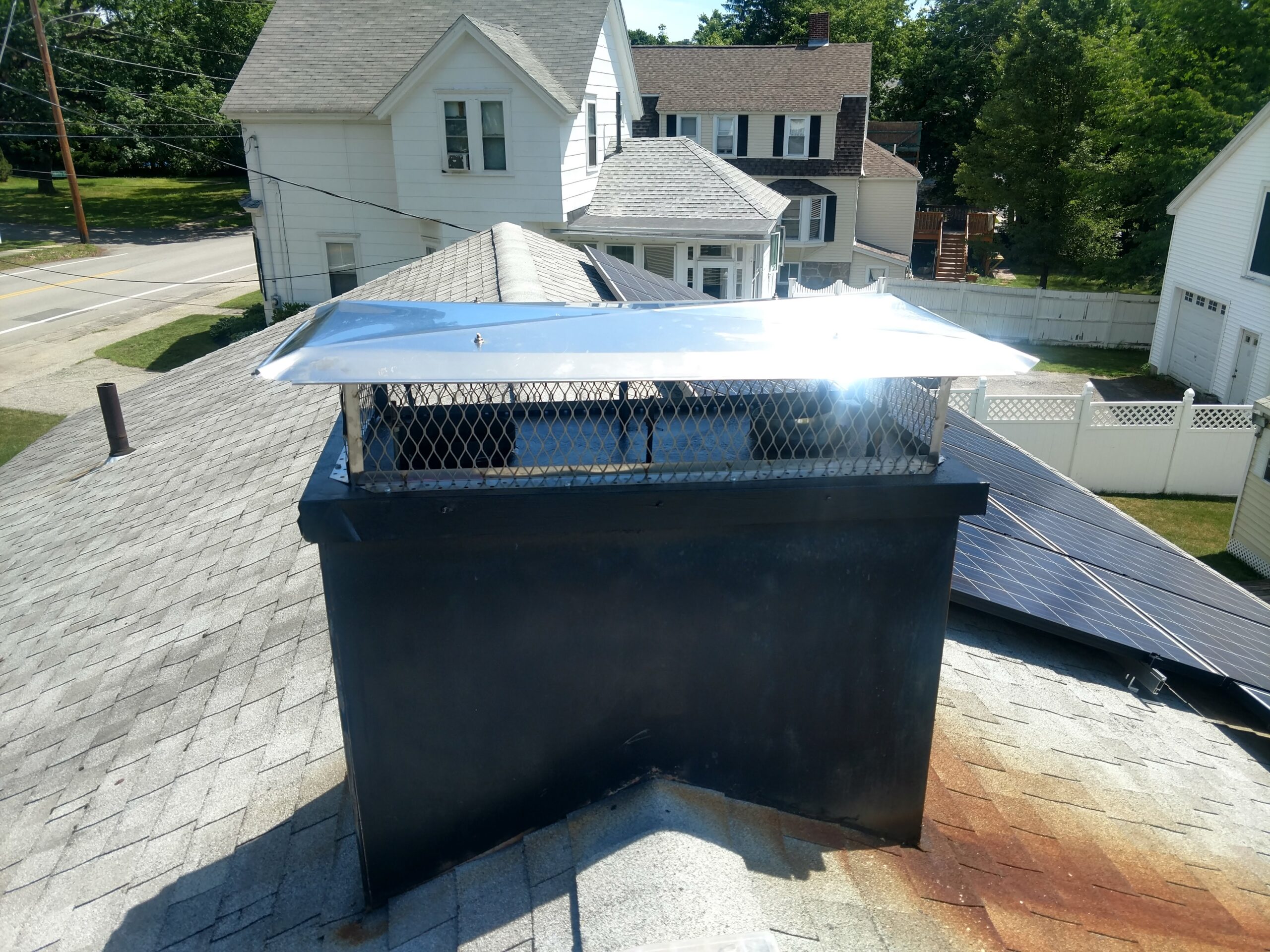 Chimney cap with a shiny metal cover sits atop a black chimney, overlooking a residential area with solar panels and nearby houses.