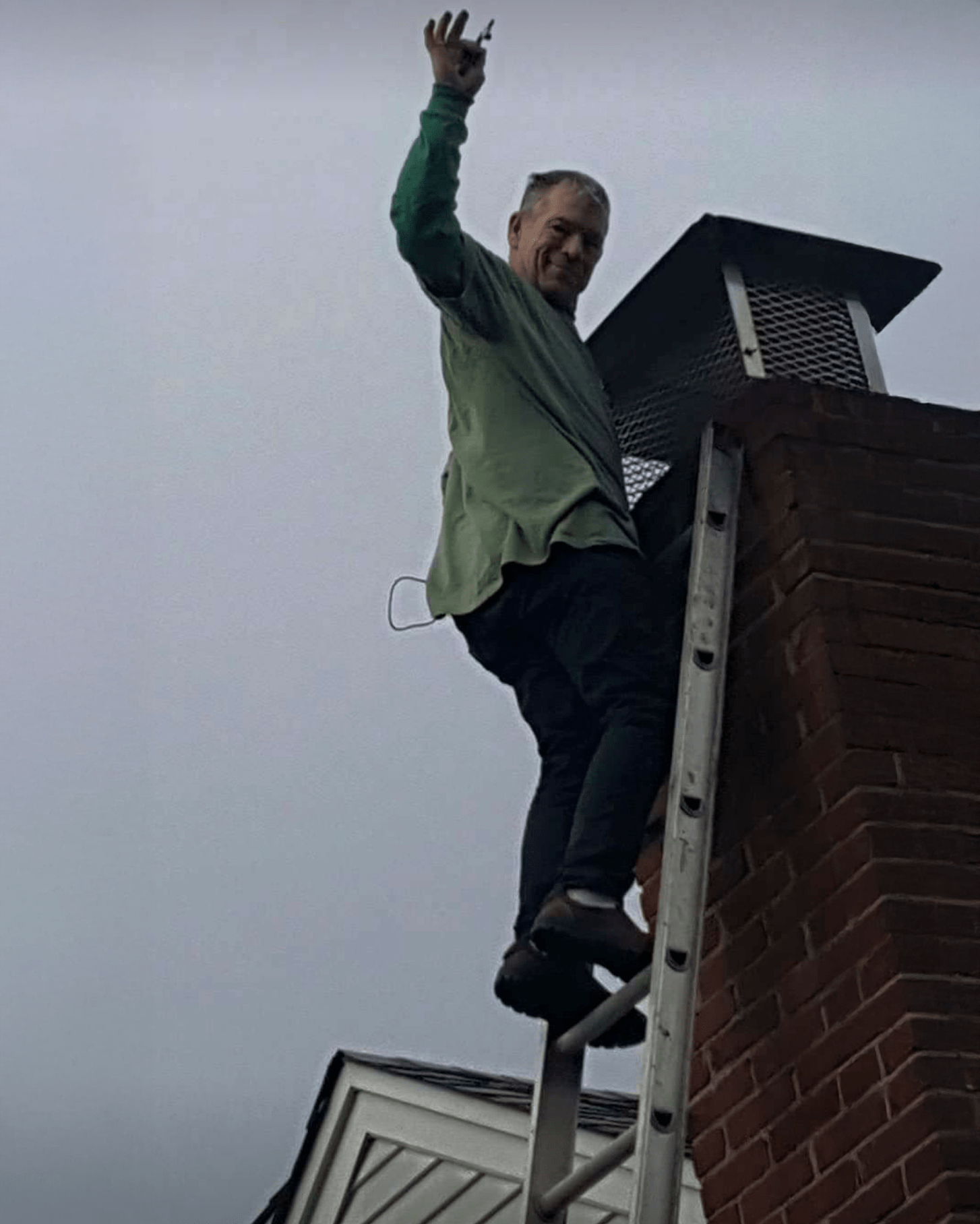 A man in a green shirt stands on a ladder next to a chimney, waving and holding a tool, during a cloudy day.
