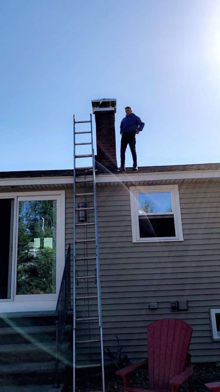 A person stands on the roof beside a chimney, with a ladder leaning against the building, under a clear blue sky.